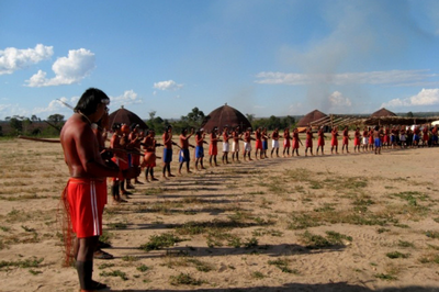 #Pracegover Foto de indígenas formando um semi círculo no centro de uma aldeia 