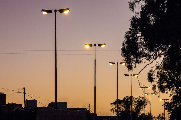 Imagem de postes de luz acesos às margens de uma rodovia no entardecer