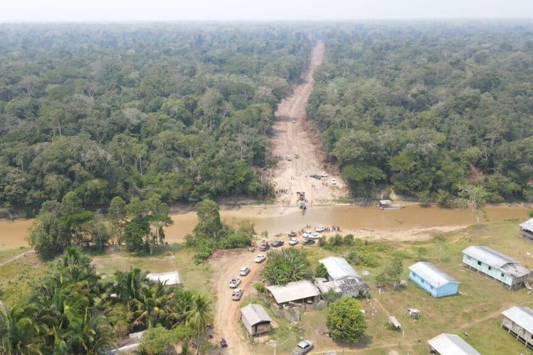 A imagem aérea mostra uma estrada de terra aberta em meio a uma vasta floresta tropical densa, conectando uma pequena comunidade rural à mata fechada. Em primeiro plano, há um aglomerado de casas modestas com telhados de zinco, cercadas por gramado, árvores frutíferas e palmeiras, onde vários veículos utilitários e carros estão estacionados ou enfileirados ao longo do caminho de terra. A estrada é interrompida por um rio de águas barrentas, onde pessoas e veículos se concentram na margem, sugerindo um ponto de travessia, antes que a via continue do outro lado como uma linha reta de terra batida que corta a floresta e desaparece no horizonte sob um céu claro e levemente enevoado.