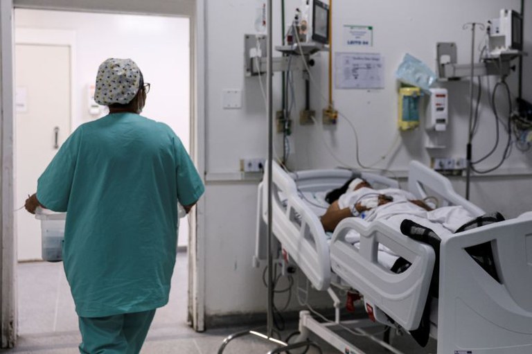 Imagem de enfermeira/ auxiliar de enfermagem passando por paciente acamado. Não é possível ver os detalhes da profissional e do(a) paciente.