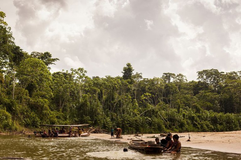 Foto mostra vegetação densa, um rio com areia e embarcações e indígenas