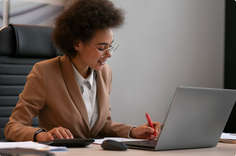 Fotografia ilustrativa mostra uma mulher negra jovem num escritório, trabalhando com um laptop, papéis e calculadora. Ela usa óculos, veste camisa branca de botão e está sentada numa cadeira de couro com espaldar alto, o que indica que ocupa um cargo de direção ou chefia.