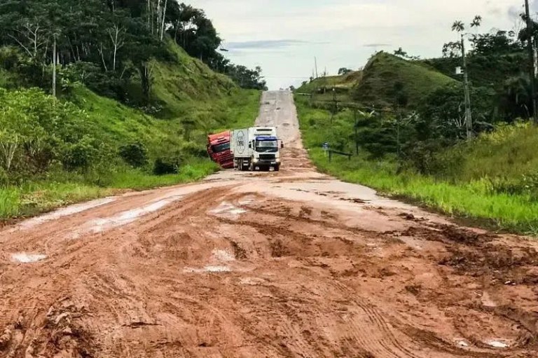 Foto mostra a rodovia com parte em terra e dois caminhões com dificuldade para passar; ao longo das duas margens vegetação