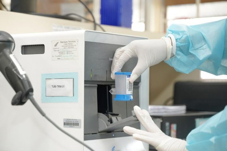 Pessoa usando avental de proteção azul e luvas insere um cartucho de teste em um equipamento de análise laboratorial.