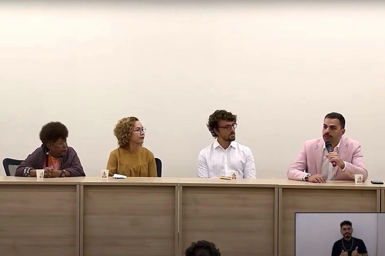 Quatro pessoas sentadas à mesa em audiência pública do Grupo de Trabalho (GT) Interministerial Memória e Verdade LGBTQIA+, em Belém do Pará. Entre as pessoas estão duas mulheres, à esquerda, e dois homens, à direita, sendo que o procurador da República República Lucas Costa Almeida Dias, mais à direita, fala ao microfone. Além deles, há um homem, intérprete de libras, no canto inferior direito da imagem.