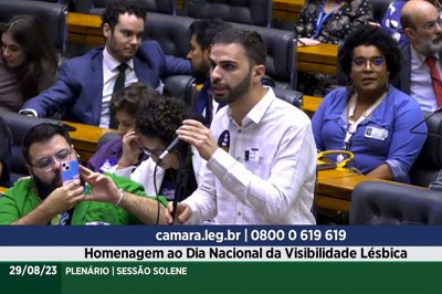 Imagem mostra um homem branco, de cabelos curtos e barba, vestindo uma camisa branca, falando ao microfone, na Câmara dos Deputados. Atrás dele, podem ser vistas algumas fileiras de cadeiras, com homens e mulheres que participam da sessão no plenário da Câmara. Sob a imagem, podem ser lidas as seguintes informações: "bamara.leg.br | 0800 0 619 619", "Homenagem ao Dia Nacional da Visibilidade Lésbica" e "29/08/2023 - Plenário | Sessão Solene".