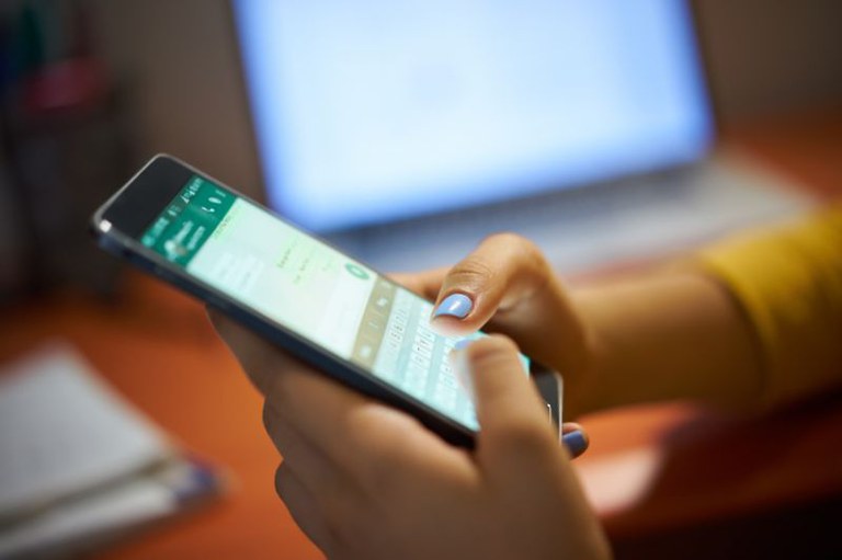 Mãos de uma pessoa com unhas pintadas de azul digitando em um smartphone com aplicativo de mensagens aberto, com notebook desfocado ao fundo.