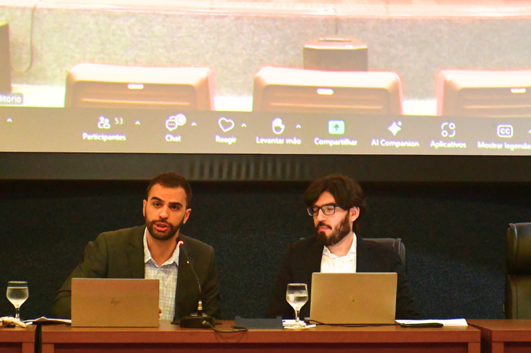 A foto anexa mostra dois homens, sentados lado a lado em uma mesa de conferência, provavelmente durante uma audiência pública ou evento formal que ocorre online, como sugerido pela tela de projeção atrás deles. O homem à esquerda, com barba escura e vestindo um blazer verde-oliva sobre uma camisa clara, está falando, com a boca aberta em meio a uma frase. O homem à direita, usando óculos e com barba e cabelo escuros, veste um terno escuro sobre uma camisa branca e olha para frente ou ligeiramente para baixo, enquanto ambos têm laptops abertos à sua frente e copos de água em cálices ao lado. A tela de projeção no fundo exibe elementos de uma interface de reunião virtual, incluindo opções como "Participantes", "Chat", "Reagir", "Levantar mão", "Compartilhar", e "Mostrar legendas", e o nome do arquivo da imagem sugere que o evento é uma Audiência Pública sobre a regulamentação do uso sacramental da Ayahuasca, realizada pelo Ministério Público Federal (MPF) no Acre.