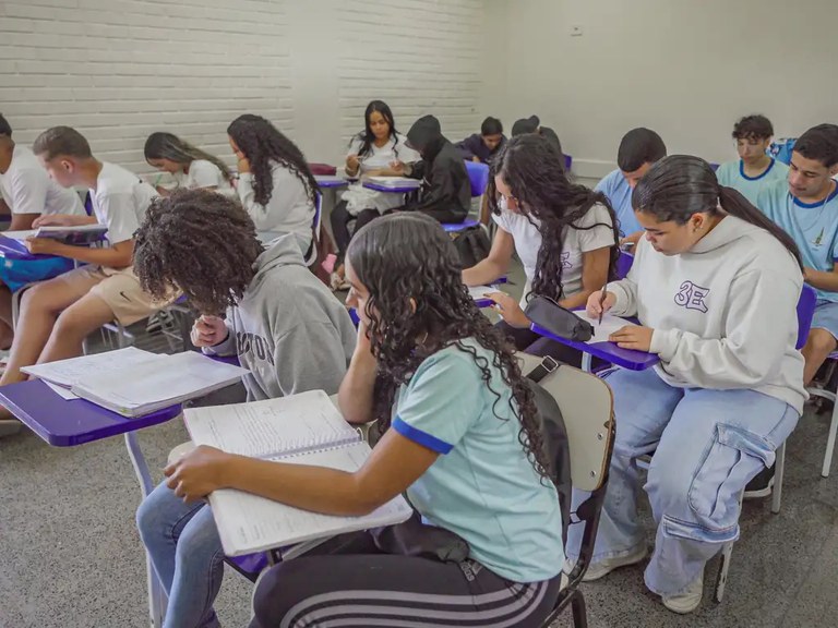 Foto de alunos do ensino médio em aula. Eles estão de cabeça baixa. As paredes da sala são brancas e as carteiras, assim como o uniforme dos alunos é na cor azul