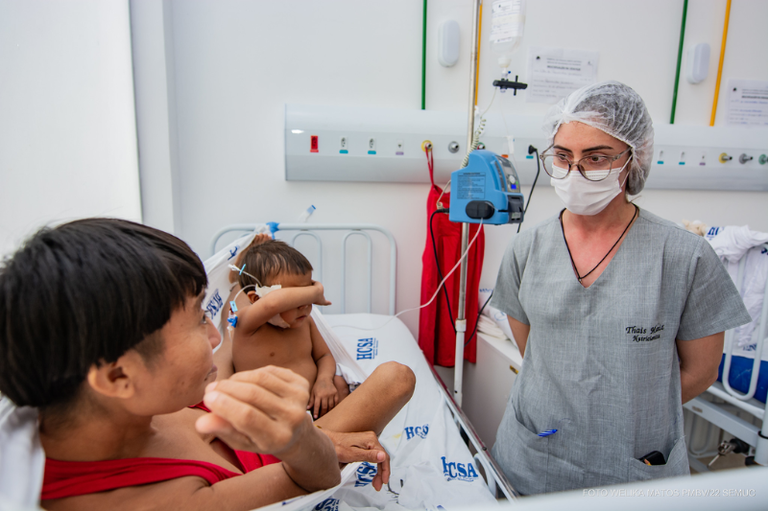 Profissional de saúde usando máscara e touca em pé ao lado de um leito hospitalar, onde uma mulher indígena e uma criança pequena estão sentadas.