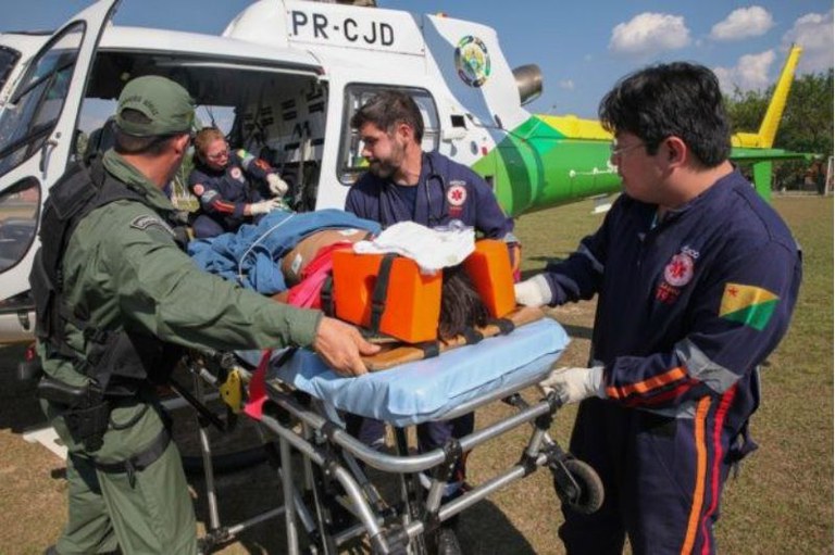 Foto mostra um paciente deitado em uma maca e quatro homens colocando a maca dentro de um helicóptero