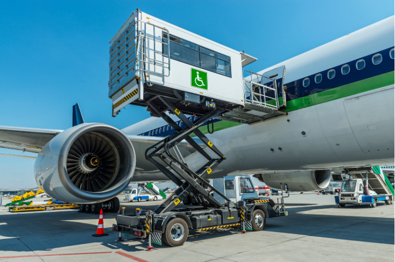 Um ambulift, veículo de assistência para pessoas com deficiência, está com sua plataforma elevada conectada à porta de um avião branco no pátio de um aeroporto. A cabine branca do elevador exibe o símbolo internacional de acessibilidade em verde. O mecanismo de tesoura que sustenta a plataforma está totalmente estendido. Ao fundo, vê-se o motor da aeronave e o céu azul límpido.