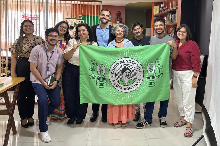Foto mostra cinco mulheres e quatro homens em pé, lado a lado em ambiente interno, posando para a foto. Uma das mulheres e um dos homens seguram uma bandeira verde à frente, com a imagem de Chico Mendes e a expressão Chico Mendes A luta continua.