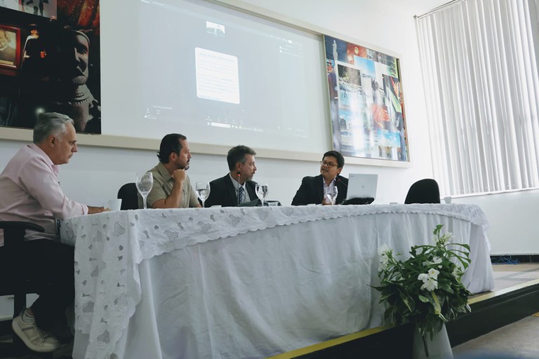 Mesa de debate realizada em auditório, com quatro participantes sentados à frente de uma longa mesa coberta por toalha branca rendada. Ao fundo, uma projeção em tela exibe uma apresentação digital, enquanto painéis fotográficos decoram a parede. Os participantes conversam durante o evento, em um ambiente institucional e acadêmico, com iluminação suave e composição documental. Na parte frontal do palco, um arranjo de flores brancas complementa a cena.