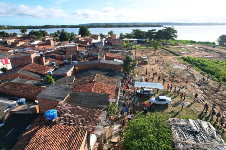 Vista aérea da comunidade do Bom Parto, com casas de tijolos aparentes e telhados de cerâmica, ao lado de uma área aberta de terra batida e vegetação. Muitas pessoas estão reunidas na parte descampada, próximas a um carro branco e uma estrutura com cobertura simples. Ao fundo, vê-se parte da Laguna Mundaú, na margem oposta, vegetação densa. O céu está parcialmente nublado e claro.