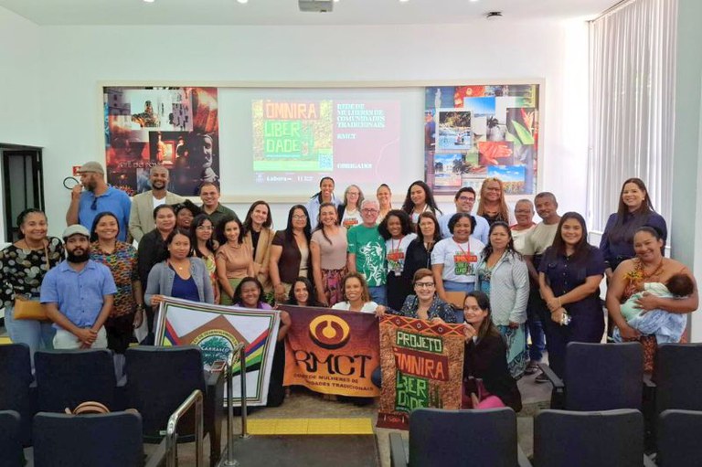 Em uma sala de auditório, um grupo diverso de cerca de 40 pessoas posa sorridente para a foto, de pé e sentadas nas primeiras fileiras. À frente, três faixas são exibidas com os nomes das organizações participantes, incluindo o projeto ÓMNIRA Liberdade e o RMCT – Rede de Mulheres de Comunidades Tradicionais. Ao fundo, um telão exibe o título “ÓMNIRA Liberdade” e informações sobre o evento. As pessoas demonstram alegria e união, em um ambiente iluminado com luz natural que entra por uma cortina lateral.