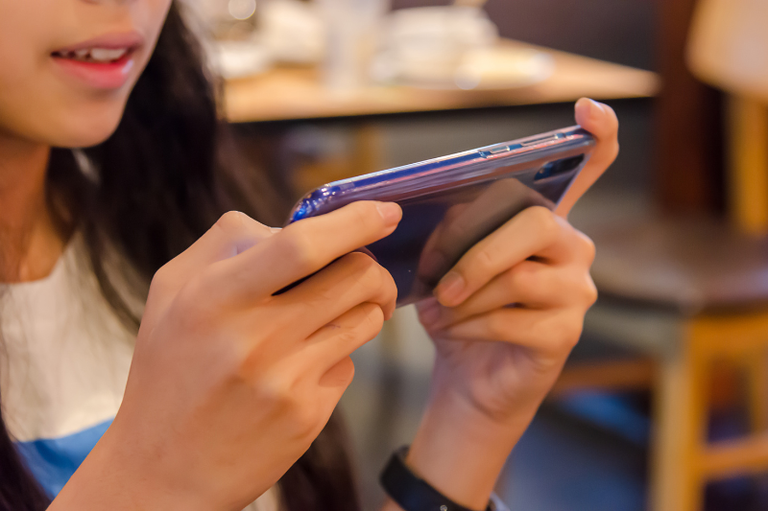 Close nas mãos de uma pessoa jovem segurando um smartphone na horizontal, focando no uso do aparelho com fundo desfocado.