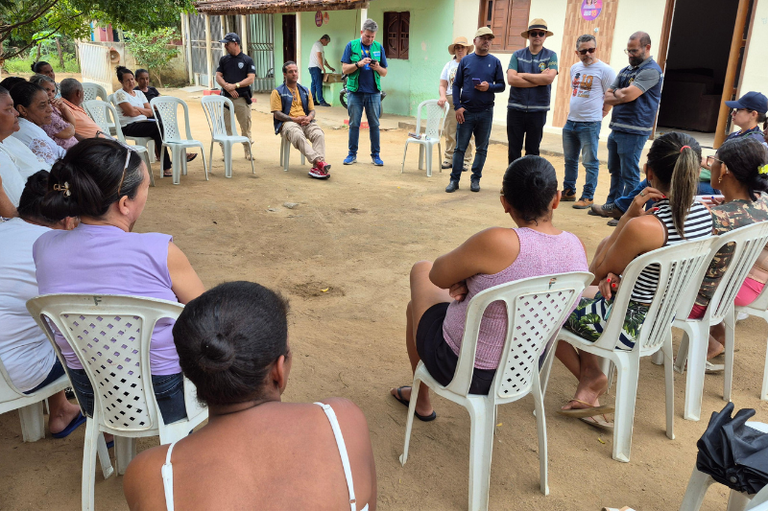 A imagem mostra uma reunião ao ar livre, em um espaço de chão batido, diante de casas simples, pintadas em tons claros de verde e bege. As pessoas estão sentadas em cadeiras plásticas brancas, formando um círculo.