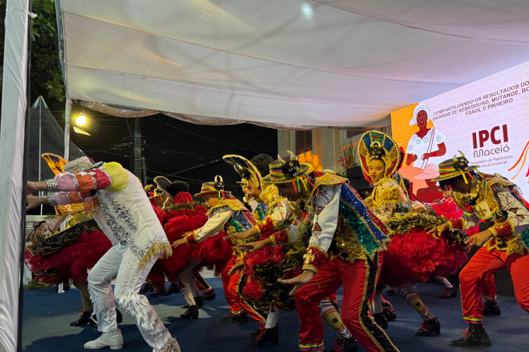 Grupo de dançarinos com trajes folclóricos coloridos e brilhantes apresentando-se em um palco sob estrutura coberta, com um painel ao fundo exibindo a marca "IPCI Maceió". 