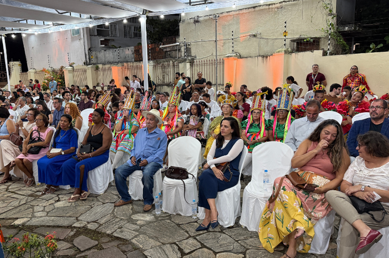 Plateia sentada em cadeiras brancas em espaço coberto, reunindo público geral e pessoas vestindo trajes folclóricos com adereços coloridos na cabeça.   