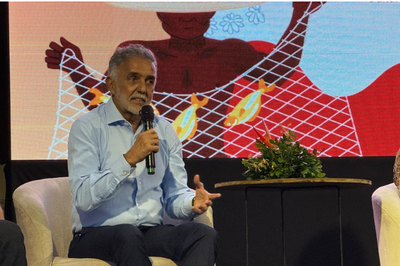 Homem de camisa social azul clara e cabelos grisalhos fala ao microfone sentado em uma poltrona branca, diante de um painel ilustrado no palco.   
