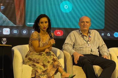 Mulher de vestido estampado fala ao microfone sentada em uma poltrona branca, ao lado de um homem, diante de um telão com transmissão de videochamada.   