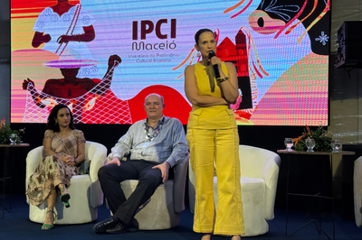 Mulher vestindo roupa amarela em pé no palco fala ao microfone, ao lado de duas pessoas sentadas em poltronas brancas. Ao fundo, um painel ilustrado exibe a marca "IPCI Maceió".   