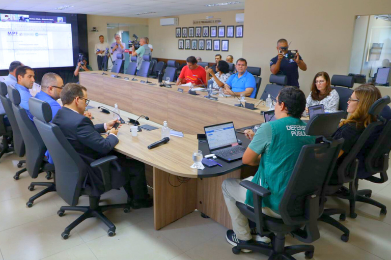 Registro fotográfico dos participantes da reunião na sala do conselho de procuradores do MPAL, diante de uma tela onde se encontram os procuradores da República, participando remotamente