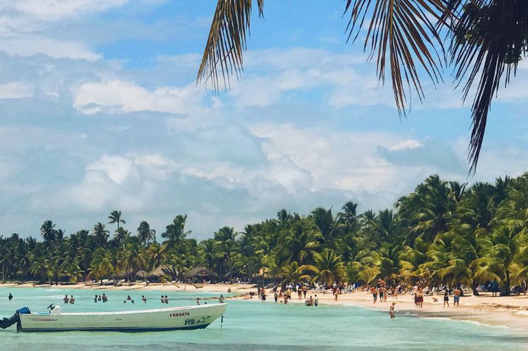 Pessoas caminham na praia com palmeiras diante da água turquesa e um barco