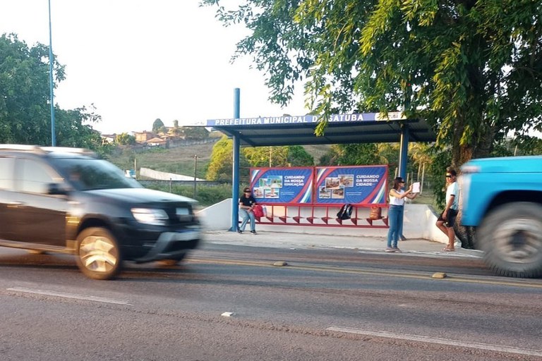 A imagem registra um momento às margens da rodovia BR-316, em frente a um ponto de ônibus da Prefeitura Municipal de Satuba, próximo ao campus do IFAL. No centro da cena, sob a estrutura metálica azul do abrigo, três jovens aguardam; uma delas está em pé segurando um cartaz branco voltado para a estrada, possivelmente pedindo carona, enquanto outra está sentada no banco e um rapaz permanece encostado em uma árvore ao lado. O ponto de ônibus exibe painéis publicitários com a frase "Cuidando da nossa educação", criando um contraste crítico com a situação de vulnerabilidade dos estudantes, que aparecem cercados pelo borrão de movimento de veículos em alta velocidade — um carro preto em primeiro plano e um caminhão azul à direita — que enfatizam o perigo e a precariedade do transporte público na região.