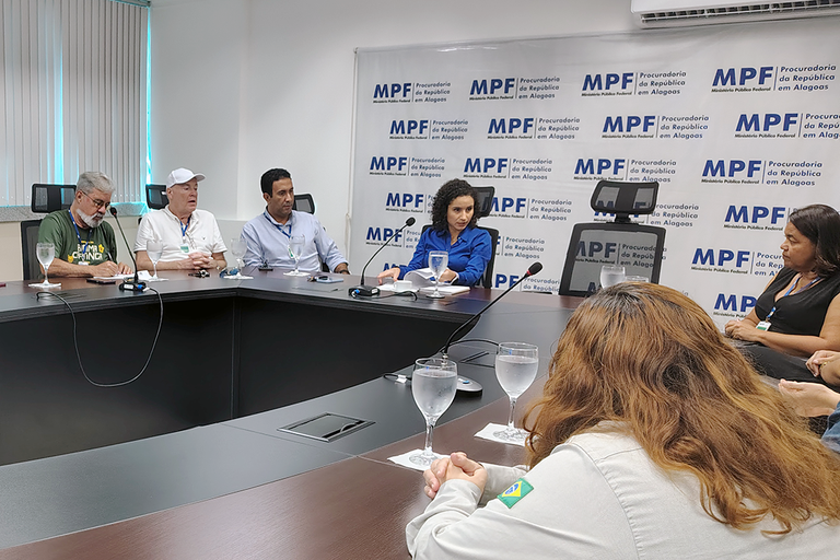 Reunião institucional em ambiente fechado, com seis pessoas sentadas em torno de uma mesa em formato de "U", onde há copos d’água, papéis e microfones. Ao fundo, observa-se uma parede branca com cortinas claras e um aparelho de ar-condicionado no alto. Em destaque, ao centro da composição, está a procuradora da República Juliana Câmara, identificada pela camisa azul — ela aparece concentrada e ligeiramente inclinada à frente. No fundo da sala, há um banner azul com a logomarca do MPF - Procuradoria da República em Alagoas. A disposição dos participantes e dos objetos sugere um momento de discussão formal sobre temas institucionais.