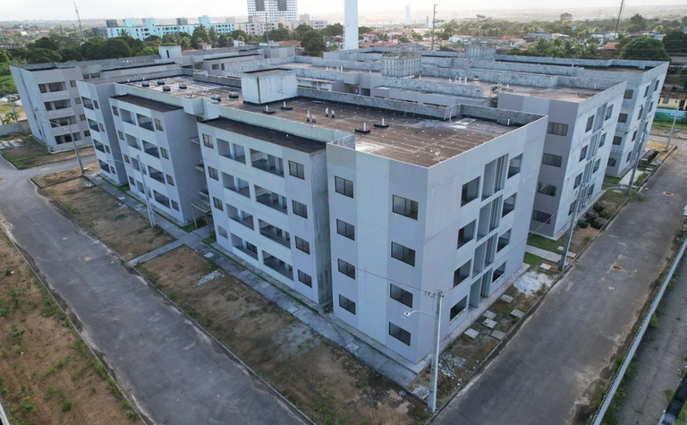 Imagem aérea, em plano aberto, de um conjunto de prédios residenciais com aparência de novos ou inacabados. São edifícios de quatro andares, com fachadas em tons de cinza claro. As janelas são retangulares, uniformes e alinhadas horizontalmente. Os prédios formam blocos simétricos, com pequenos recuos e entradas individuais. Não há varandas visíveis.