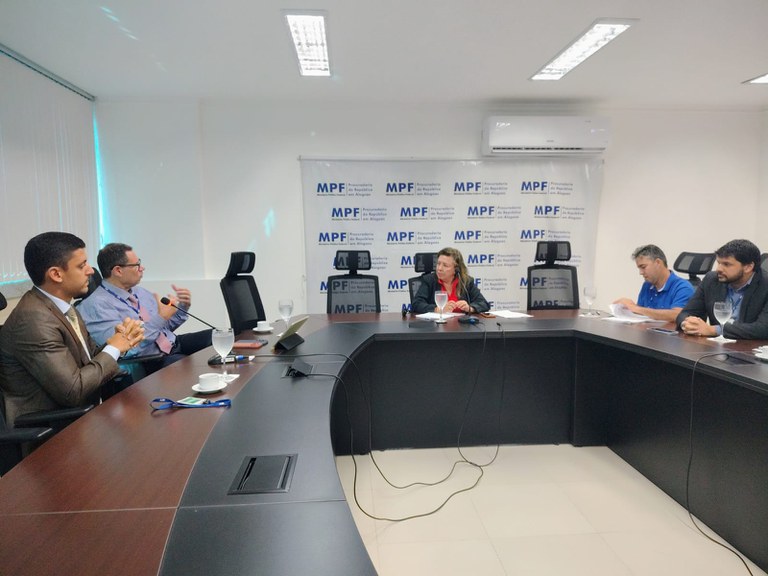 Reunião realizada em uma sala do Ministério Público Federal em Alagoas. Ao centro, sentada à mesa, está a procuradora da República Niedja Kaspary. À esquerda e à direita, estão representantes da Associação dos Despachantes Documentalistas e dos Conselhos Regional e Federal de Despachantes Documentalistas. Os participantes estão sentados ao redor de uma mesa de reuniões, diante de um painel com a identificação do MPF.
