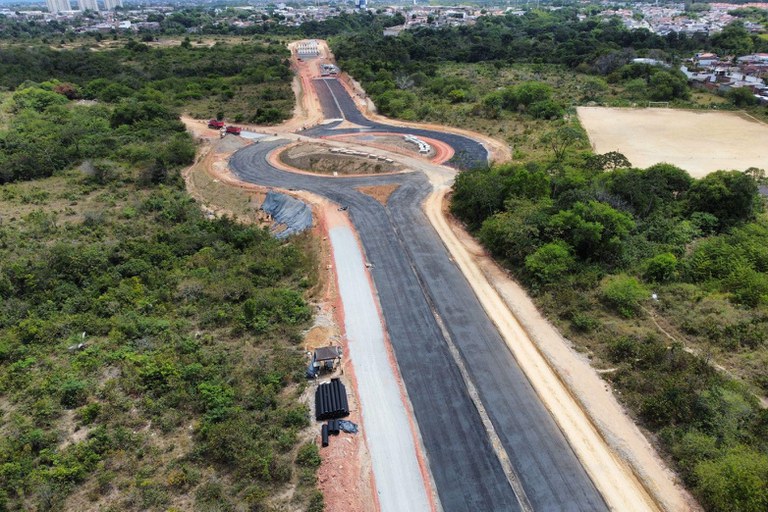 A imagem apresenta uma vista aérea de um canteiro de obras de infraestrutura viária em Maceió, Alagoas, destacando a construção de um trecho da Via Linha Verde. No centro da composição, observa-se uma rotatória em processo de pavimentação, conectando vias de asfalto escuro que contrastam com o solo de terra avermelhada e as faixas laterais de cascalho claro. A obra está inserida em uma vasta área de vegetação nativa e densa, com maquinários e materiais de construção, como tubulações pretas e caminhões, visíveis ao longo do traçado. Ao fundo, a linha do horizonte revela a transição entre a zona de preservação ambiental e a malha urbana da cidade sob um céu claro.
