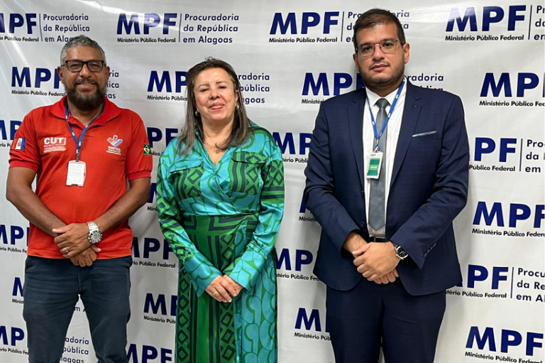 Na imagem, três pessoas posam para uma foto em frente a um painel institucional do Ministério Público Federal (MPF), com o texto “Procuradoria da República em Alagoas”.