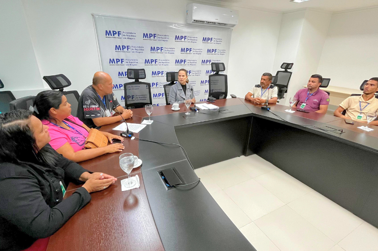 Em uma sala de reuniões do Ministério Público Federal (MPF), oito pessoas estão sentadas ao redor de uma mesa em formato de “U”. Ao centro, na cabeceira da mesa, está uma mulher de terno cinza, aparentemente conduzindo a reunião. À esquerda da imagem, quatro pessoas — três mulheres e um homem — estão sentadas, vestindo roupas casuais e com crachás no pescoço. À direita, outros três homens também estão sentados, igualmente com crachás visíveis. A parede ao fundo exibe um painel institucional do MPF, com o nome da Procuradoria da República em Alagoas repetido sobre fundo branco. Há copos de vidro sobre a mesa e aparelhos de microfone embutidos. O ambiente é claro, com piso branco e ar-condicionado fixado na parede.