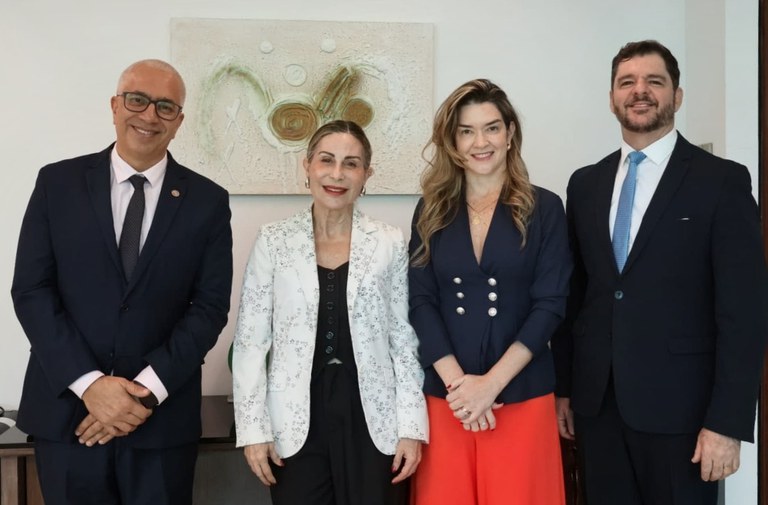 Quatro pessoas posam lado a lado para fotografia em ambiente interno. Da esquerda para a direita, Carlos Henrique Macedo, de terno escuro e gravata; Raquel de Melo Teixeira, de blazer branco estampado sobre roupa preta; Bruna Rizzato Barbosa, de blazer azul-marinho e calça vermelha; e Ronaldo Guilherme Campos, de terno escuro e gravata azul. Ao fundo, há uma parede clara com uma obra de arte em tons neutros.