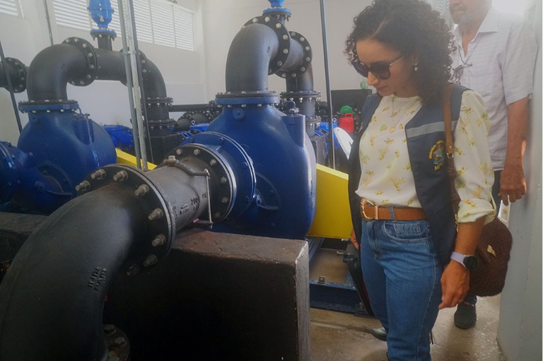 Uma mulher usa colete institucional e óculos escuros enquanto observa um conjunto de grandes tubulações e válvulas industriais de cor azul, instaladas em um ambiente fechado. Ela está de pé ao lado dos equipamentos, olhando atentamente para as estruturas metálicas, que aparentam fazer parte de um sistema hidráulico ou de bombeamento. Ao fundo, outra pessoa acompanha a visita técnica.