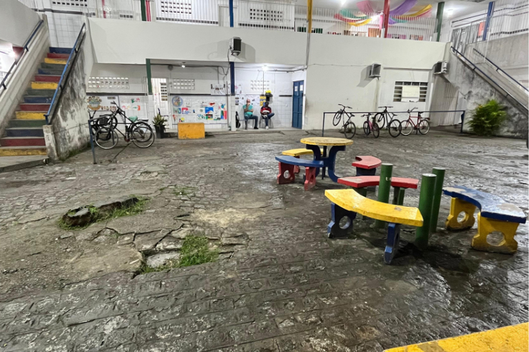 Foto tirada na escola onde mostra uma mesa infantil no meio de um solo desgastado de concreto. O ambiente aparenta pouca qualidade.