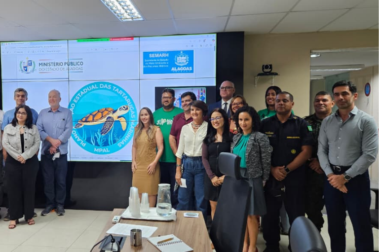 Na imagem, um grupo de pessoas posa para foto em uma sala de reuniões. Ao fundo, há uma tela grande com os logotipos do Ministério Público do Estado de Alagoas, da Secretaria de Estado do Meio Ambiente e Recursos Hídricos (Semarh/AL) e do Plano Estadual das Tartarugas Marinhas.