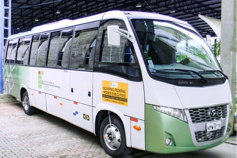 Ônibus escolar branco e verde do Instituto Federal Fluminense (IFF) estacionado em um pátio coberto, com identificação do Governo Federal na lateral.