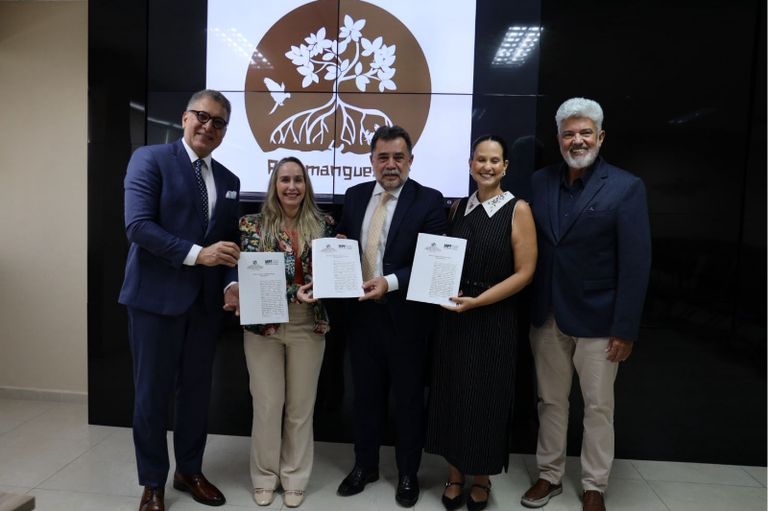 Cinco pessoas posam lado a lado em um ambiente institucional, segurando documentos. Ao fundo, há um painel com a imagem de uma árvore estilizada e a palavra “manguezal”. Três delas exibem papéis assinados.