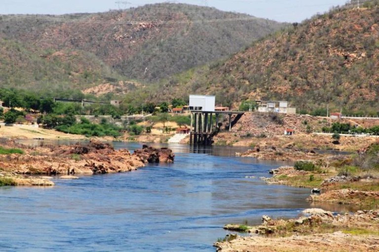 Vista ampla de um rio de águas calmas que atravessa uma paisagem semiárida, com margens rochosas e vegetação esparsa. Ao fundo, há um conjunto de morros cobertos por mata seca. No centro da imagem, uma estrutura elevada com pilares sustenta uma edificação branca sobre o rio, provavelmente parte de uma barragem ou estação de captação. À direita, há pequenas construções e uma estrada próxima à margem. O céu está limpo e azul.
