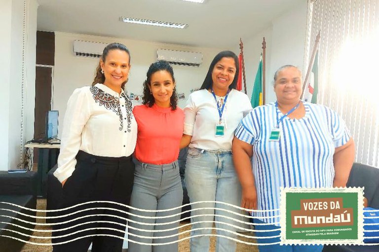 A foto mostra quatro mulheres posando lado a lado, sorrindo para a câmera em uma sala institucional. Todas usam crachás de identificação. Ao fundo, aparecem bandeiras, ar-condicionados e parte do mobiliário do ambiente.