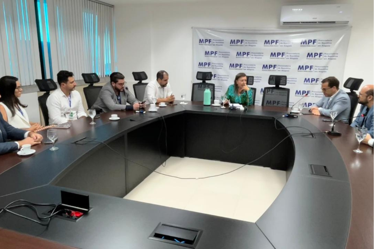A imagem mostra uma reunião realizada em uma sala de conferências da Procuradoria da República em Alagoas (MPF/AL). Os participantes estão sentados em torno de uma grande mesa em formato de “U”, com notebooks, copos e xícaras à frente.