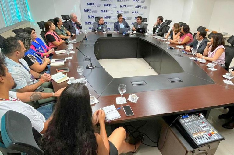 Em uma sala de reuniões, cerca de 15 pessoas estão sentadas ao redor de uma grande mesa oval. Elas conversam e tomam notas, com copos de água e xícaras à frente. No fundo, há um painel com a logomarca do MPF.