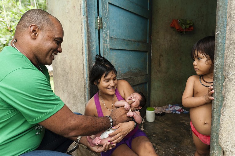 Interação entre um profissional de saúde, uma mãe indígena e seu bebê na entrada de uma casa rústica de barro com porta azul, enquanto uma criança observa.
