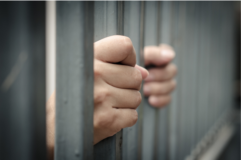 A imagem mostra uma pessoa segurando as grades de uma prisão ou de uma cela. As mãos estão em primeiro plano, agarrando firmemente as barras verticais. O foco está nas mãos, enquanto o restante da imagem, incluindo o fundo, está desfocado. A cena sugere um sentimento de confinamento e desespero.