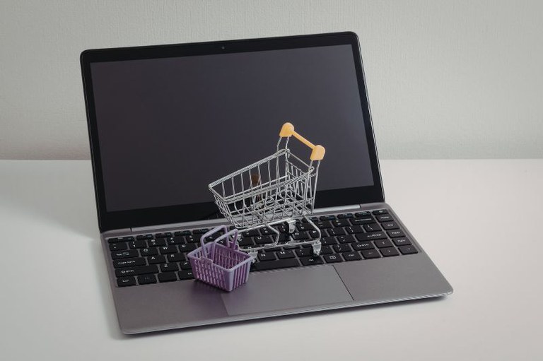 Foto mostra um notebook aberto e miniaturas de um carrinho de compras e de uma cesta vazias por cima do teclado