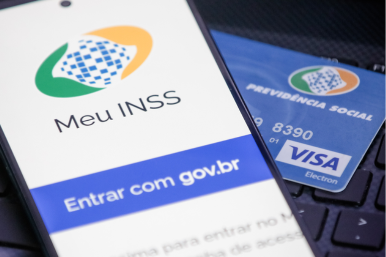 Foto mostra um cartão magnético azul da Previdência Social sobre um teclado preto de computador; sobre metade do cartão, há um telefone celular com a tela de início do Meu INSS